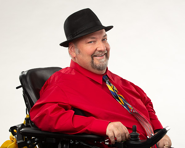 Dave in his wheelchair wearing a red shirt, multicolored tie and black hat
